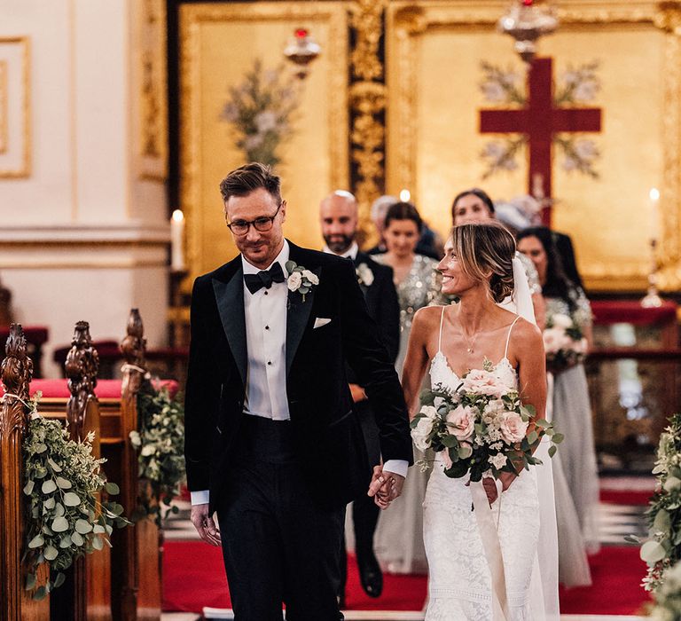 The bride and groom walk holding hands back down the aisle with the groomsmen and bridesmaids in sparkly grey dresses behind them 