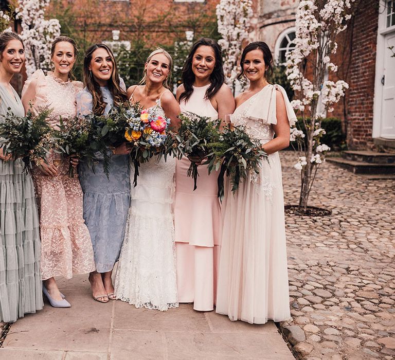 The bride stands smiling with the bridal party wearing mismatched bridesmaid dresses in different shades and patterns for fun greenery theme wedding