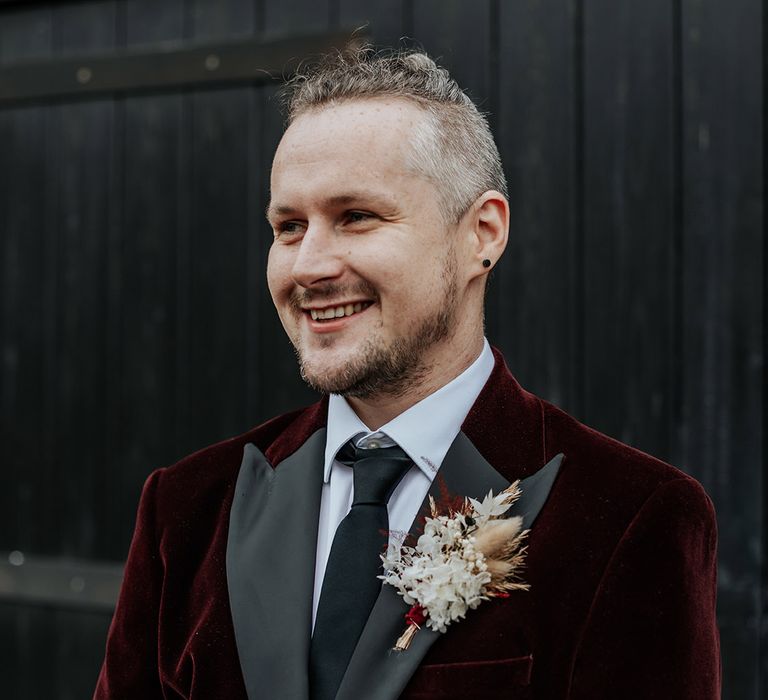 The groom wears a burgundy velvet suit jacket with a dried flower buttonhole and a black tie 