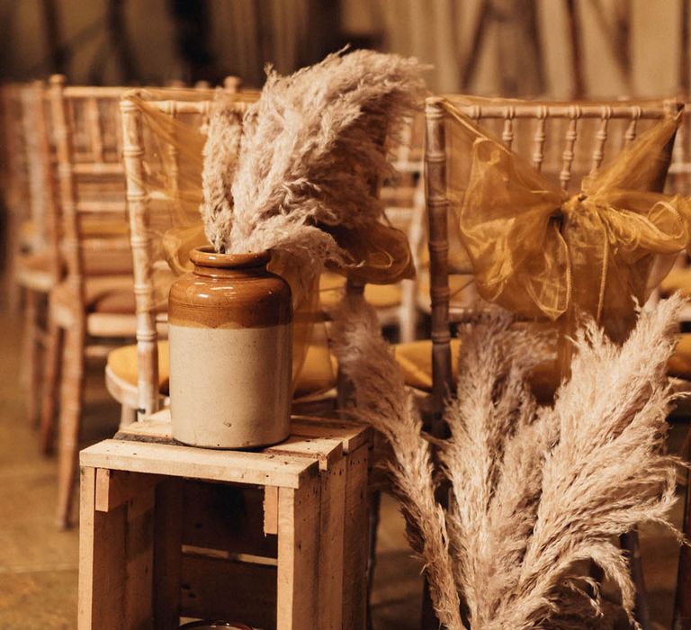 Pampas grass in two tone vases and wooden crates to decorate the aisle with wedding chairs decorated with sheer yellow bow 