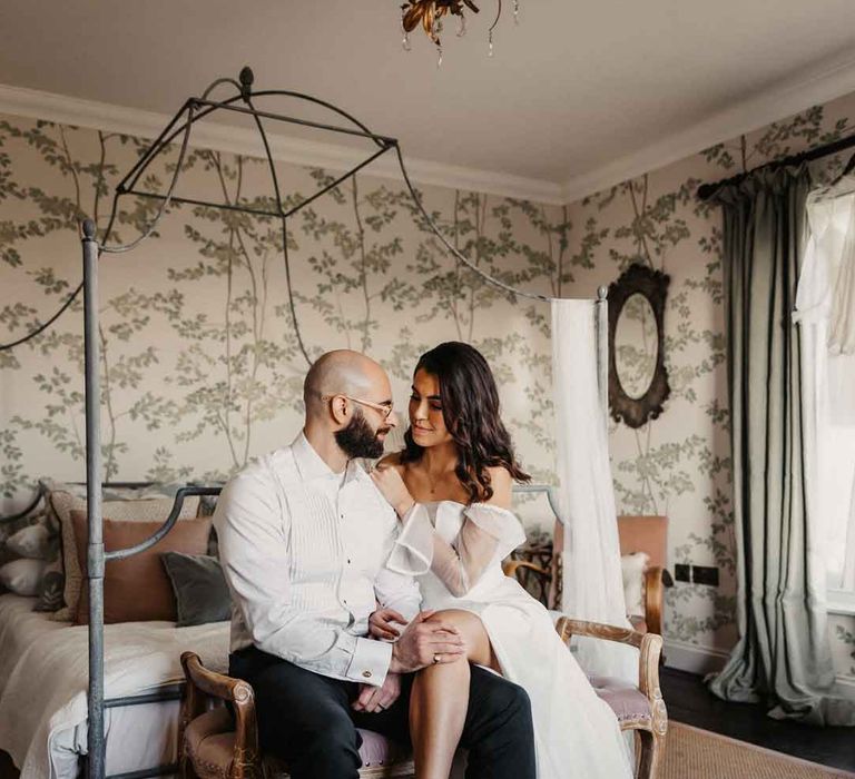 Bride in sheer puff sleeve wedding dress with front slit sitting with groom in white shirt and black suit trousers 