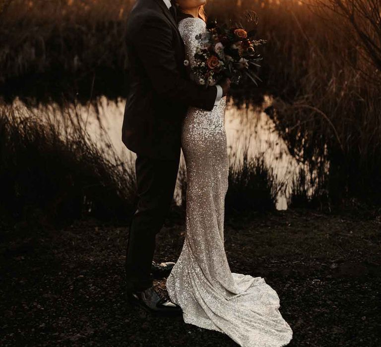 Bride in silver sparkly wedding dress embracing groom in classic black tuxedo doing golden hour couples portraits at Elmley Nature Reserve wedding venue 