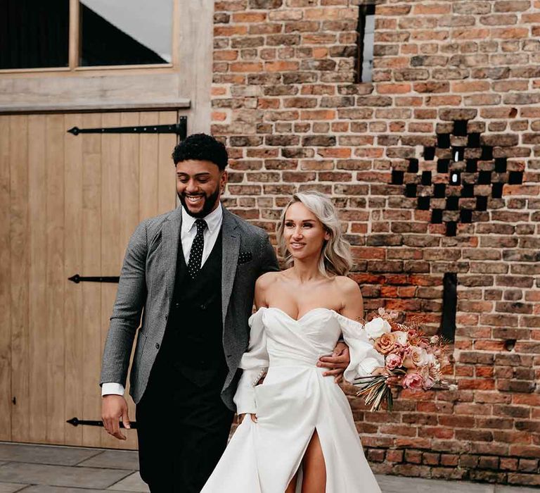 Bride in off the shoulder long sleeve wedding dress with slight cowl neck and front leg slit holding blush pink dried flower bridal bouquet walking with groom in grey blazer, black waistcoat and navy polkadot tie at Lodge Farm