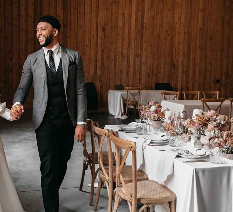 Bride in off the shoulder long sleeve Nori Ivy wedding dress with slight cowl neck and front leg slit walking with groom in grey blazer, black waistcoat and navy polkadot tie