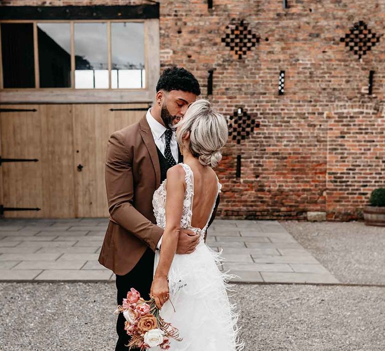 Groom in mocha brown blazer, black waistcoat, polka dot tie and pocket square and dried blush boutonniere hugging bride in corset top sheer floral 3d appliqué wedding dress with fringe overlay 