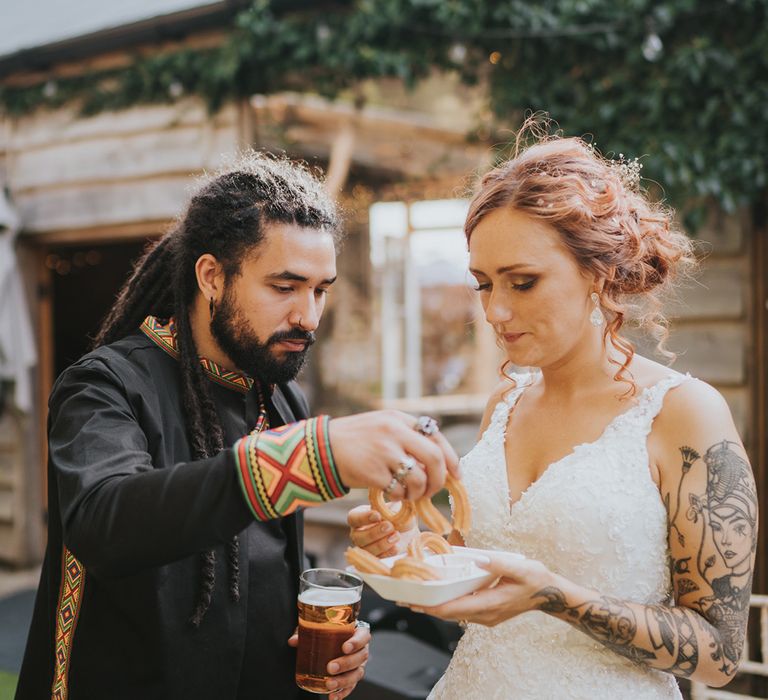 bride in a lace wedding dress and groom in a black Caribbean outfit eating churros at their wedding