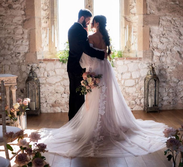 Bride in puddle train ethereal off the shoulder wedding dress with detachable puff tulle sleeves and embroidered rose 3d appliqué details embracing groom in classic black tux at Pentney Abbey wedding venue 