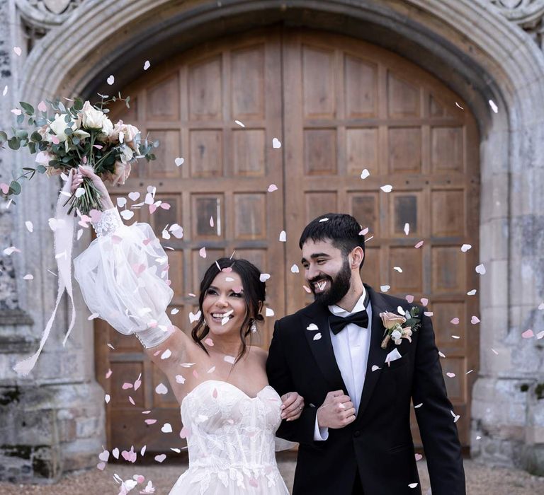 Bride in front leg slit ethereal off the shoulder wedding dress with detachable puff tulle sleeves and embroidered rose 3d appliqué details and groom in classic black tuxedo doing colourful confetti exit at Pentney Abbey wedding  