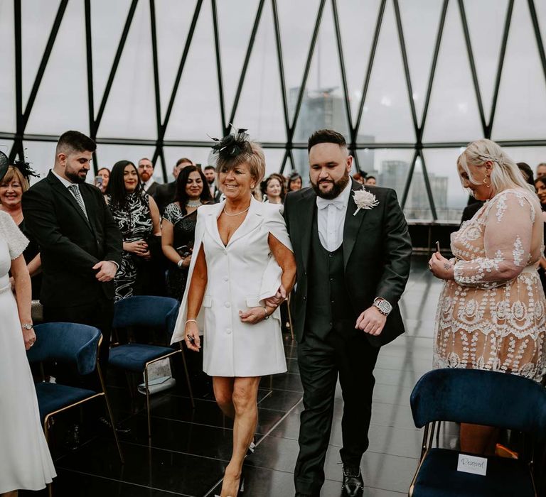 Mother of the groom wearing short white blazer dress with cut out sleeves and black wedding fascinator walking groom in three piece black tuxedo with off white bow tie, white garden rose, lavender twig and dried foliage boutonniere and grooms Louboutins shoes down the aisle of The Gherkin wedding venue 