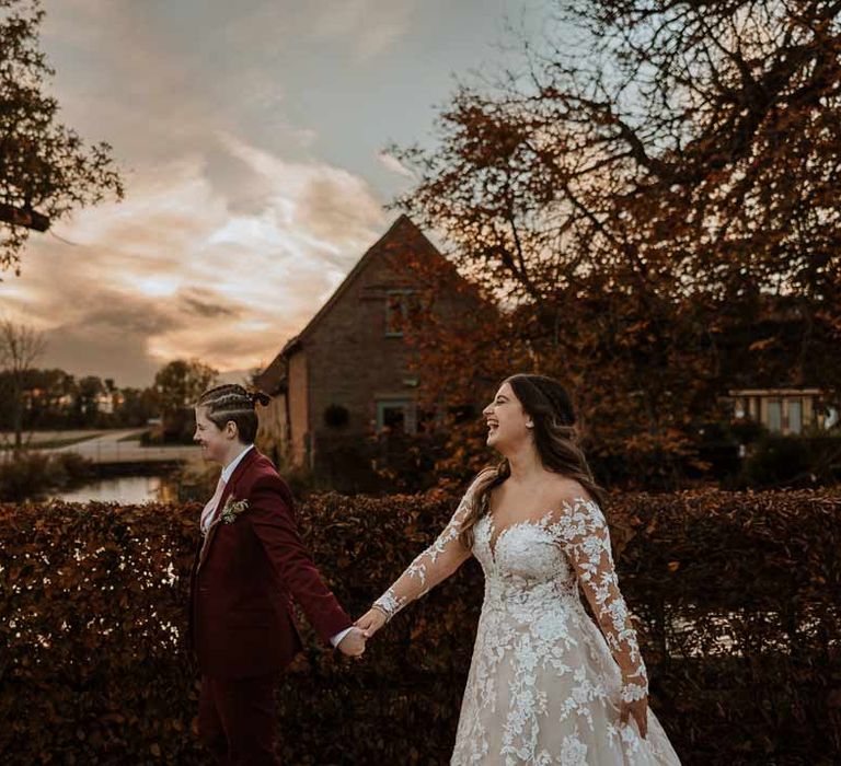 Bride in maroon bridal suit with grey waistcoat, dusky pink tie and autumnal dried flower boutonniere walking hand in hand with bride in long sleeve lace illusion sleeve wedding dress with overlay