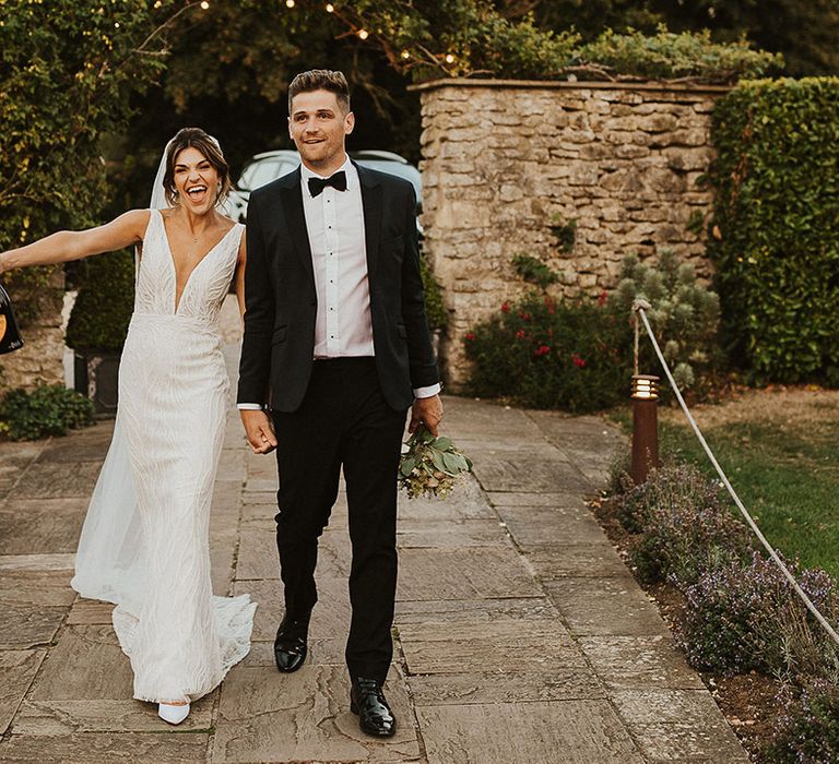 Bride in fitted Made With Love wedding dress walking along with champagne walking along with groom in black tie 