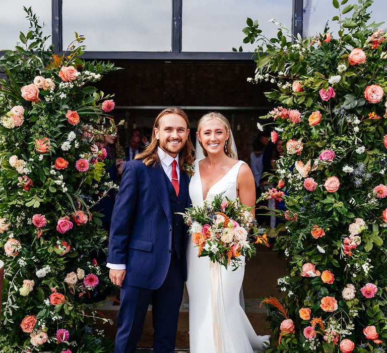 Colourful flower column arrangements with the groom wearing a blue three piece suit and bride in a Savin London wedding dress 