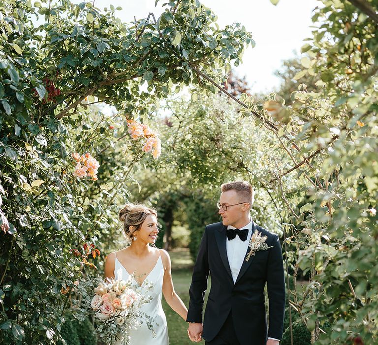 Bride in a slip dress for wedding with colourful pastel bouquet walking along with groom in black tie