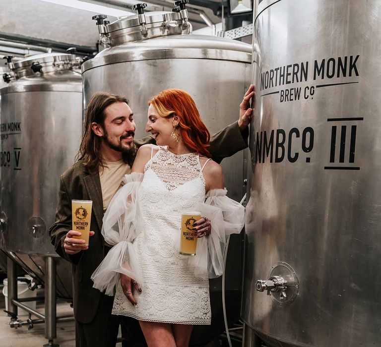 Bride wearing 70s-inspired boatneck short lace wedding dress with detachable tulle puff sleeves and platform gold wedding heels standing by beer fermenter with groom wearing deep brown suit with off-white t-shirt and vans at Northern Monk Refectory Leeds