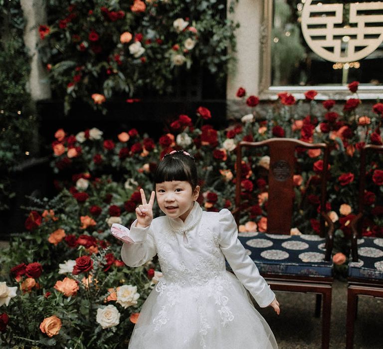 The bride and groom's daughter in a princess style white dress and pearl necklace giving a peace sign 