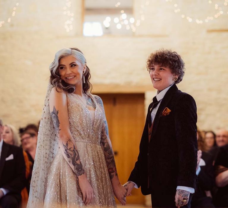 Bride in dark velvet bridal jacket with rust waistcoat and pocket square, black suit trousers and gold pocket watch holding hands with bride in sleeveless deep v-neck sparkly gold wedding dress with sheer and gold polka dot wedding veil at the alter of Kingscote Barn Tetbury 