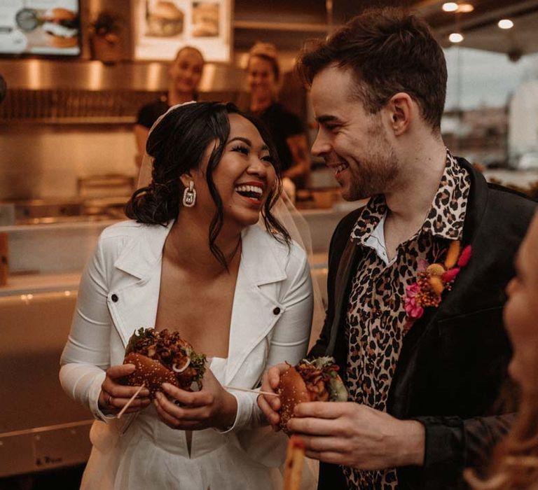 Bride in white leather jacket having wedding burger with groom in wet look black grooms suit with leopard print shirt and pink and orange boutonniere