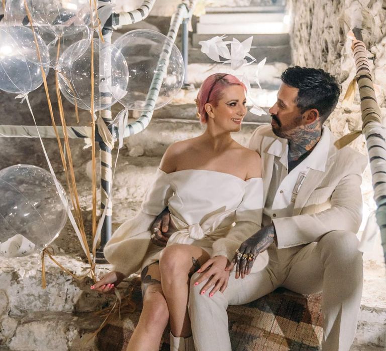 Bride in alternative off the shoulder puff sleeve short wedding dress and white chunky wedding boots sitting with groom in beige linen suit and embroidered linen shirt on the steps at Margate Caves