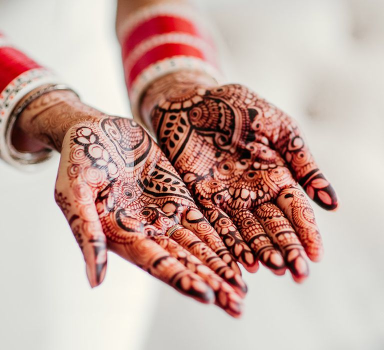 Bride holding out her hands to show off her detailed intricate wedding henna designs with red bangles 