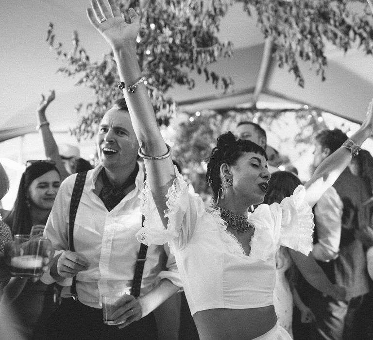 Bride in ruffle bridal separates dances with hands in the air surrounded by wedding guests at tipi wedding