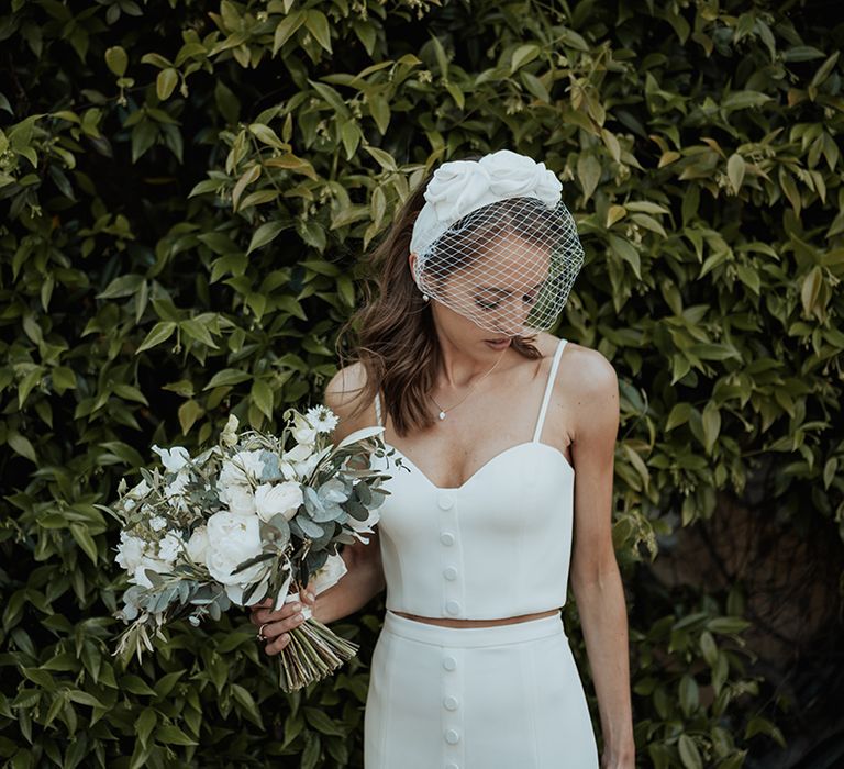 bride in separates with headband and blush veil holding a white flower and green foliage wedding bouquet 