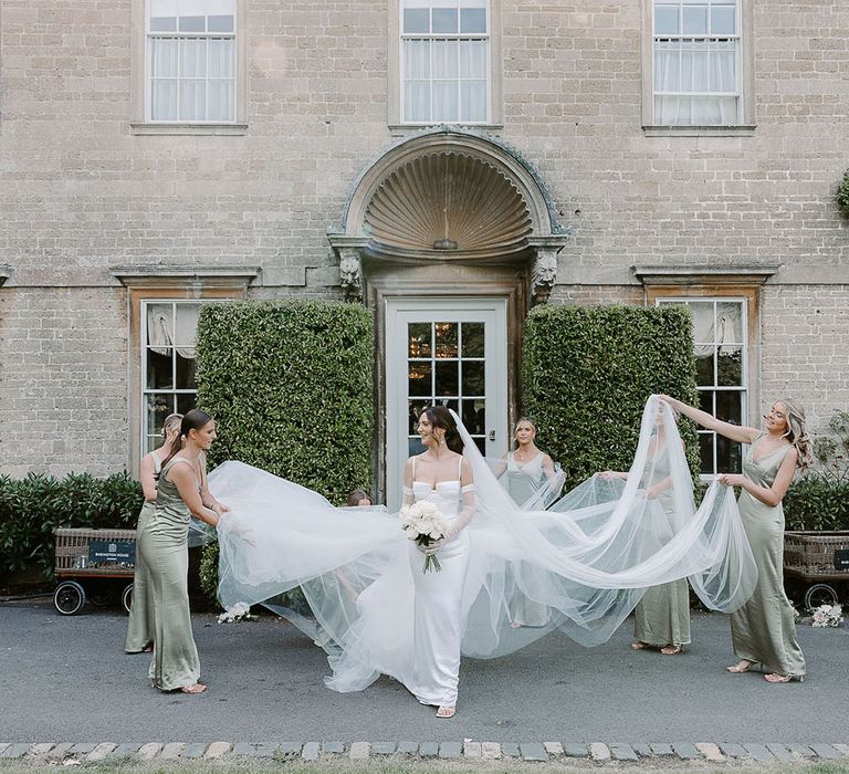 Bridal party in green satin bridesmaid dresses holding her veil and overskirt 