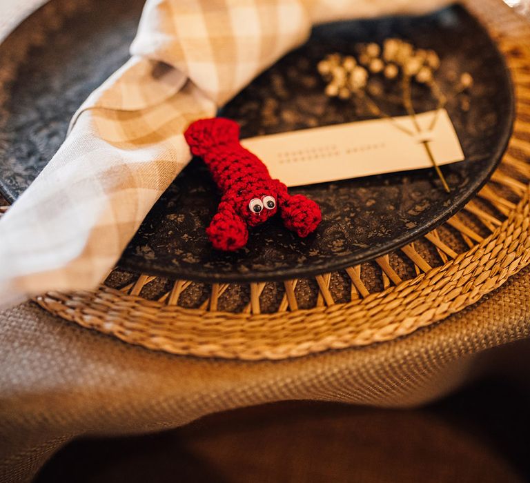 Wedding place setting with wicker place mat, black plates, crochet red lobster favours, wedding menu and place card with checkered napkin