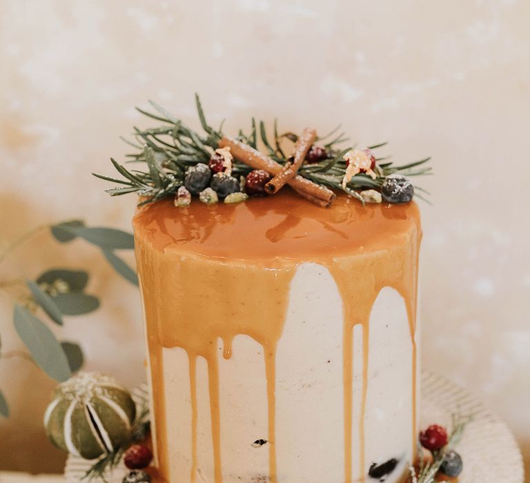 Single tier tall wedding cake covered in white frosting and drip wedding icing topped with cinnamon sticks and evergreen pieces 