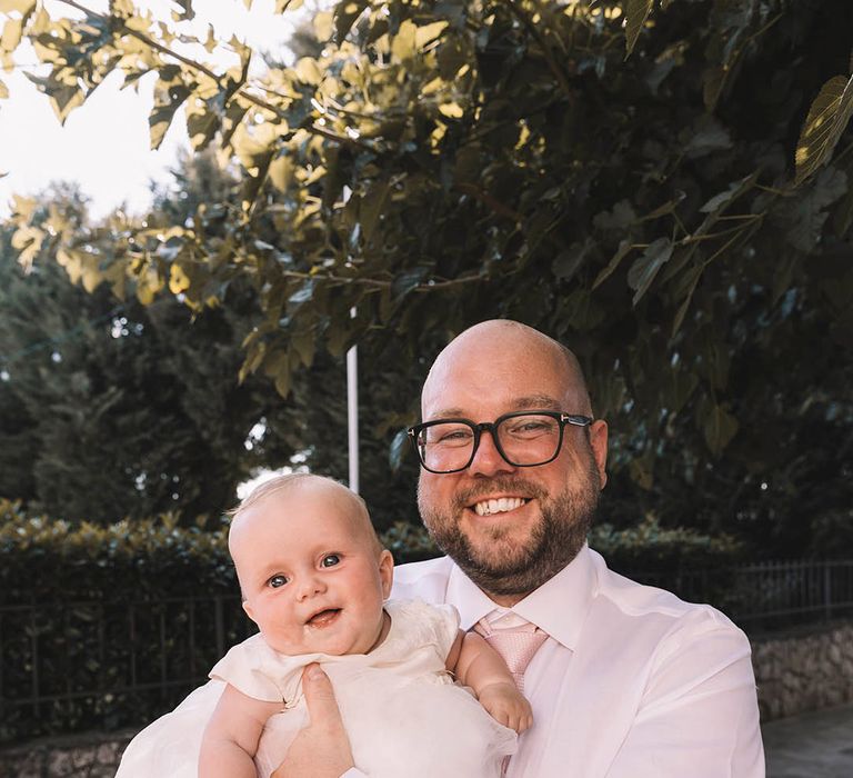 Groom in white shirt with pink tie holding baby 