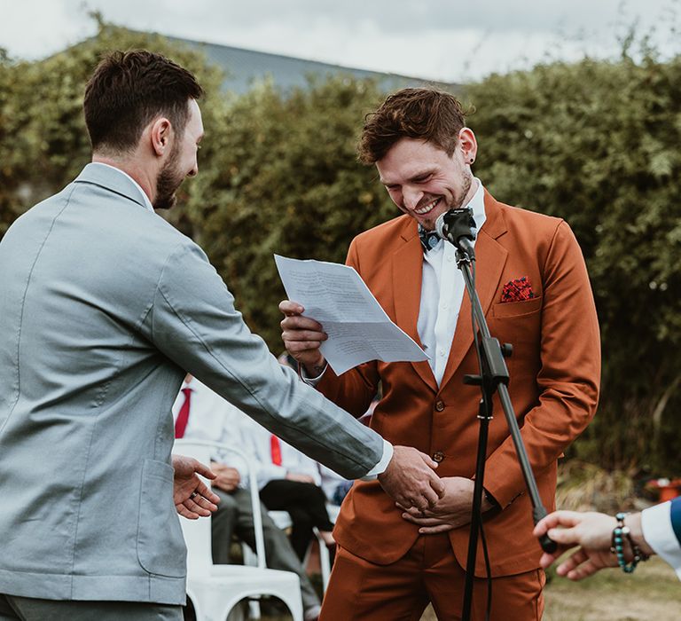 Groom in a grey wedding suit with groom in a burnt orange suit exchange their vows at same sex ceremony 