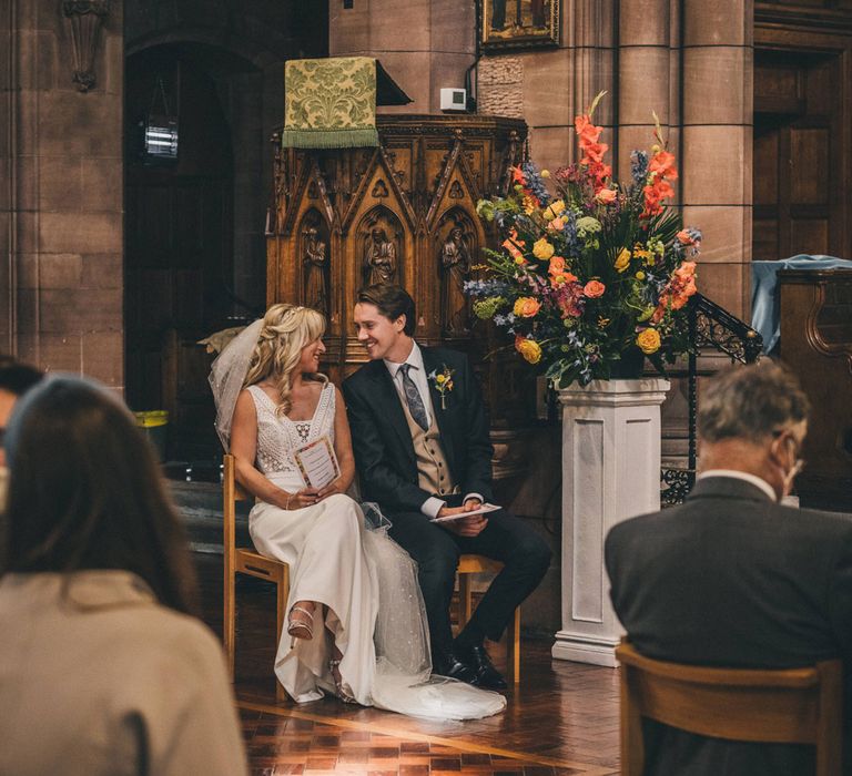 Bride sitting in floor-length veil and Justin Alexander wedding dress smiles at groom sat next to her in church wedding ceremony 