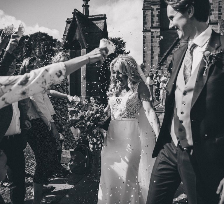 Laughing bride in Justin Alexander wedding dress holding colourful bouquet holds hand with groom whilst wedding guests throw confetti outside of church