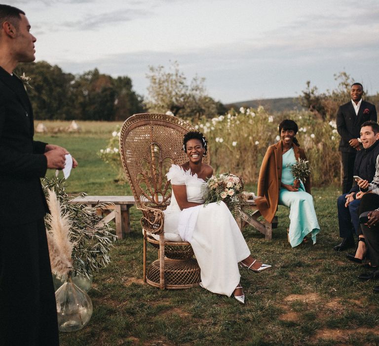 Outdoor wedding ceremony with West African bride in an off the shoulder wedding dress and silver shoes sitting on a wicker peacock chair 