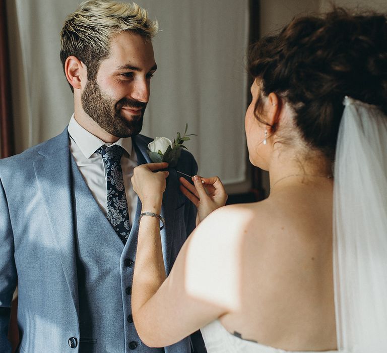 Bride in a strapless wedding dress with a cathedral length veil putting on her grooms white rose buttonhole 