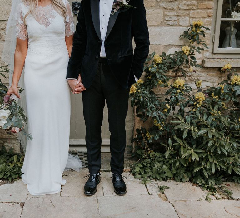 Bride in white Charlie Brear dress, white lace Augusta Jones top and full length veil holding hands with groom in black velvet Hugo Boss suit in the grounds at Caswell House wedding