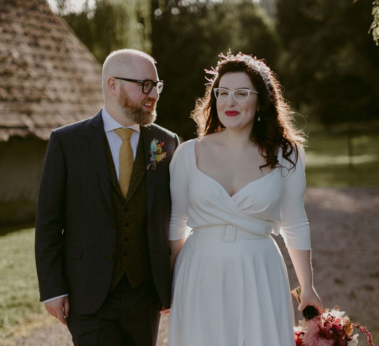 Groom in navy suit and yellow tie stands with bride in white cat eye glasses and bridal headband holding white and pink rose and pampas grass bouquet