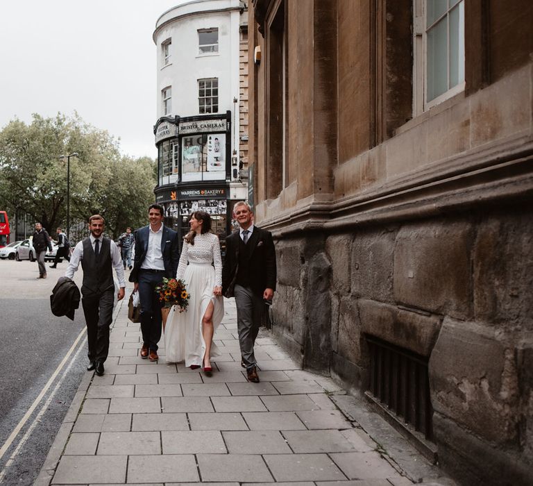 Bride in long sleeved lace Self Portrait wedding dress and groom in brown woollen suit walk through streets of Bristol holding hands alongside two friends