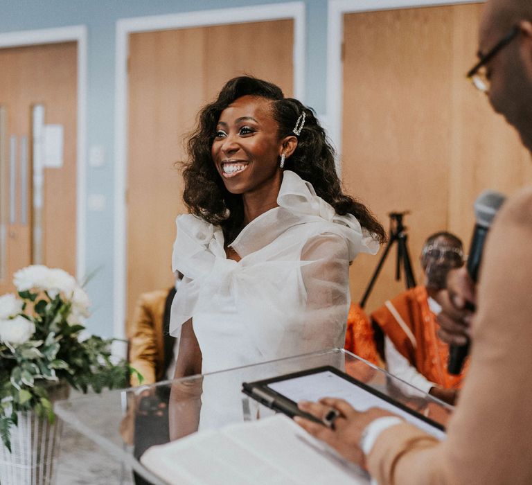 Smiling bride with curled wedding hair and white Bec + Bridge wedding dress with shoulder bows stands at the altar with groom in grey suit jacket