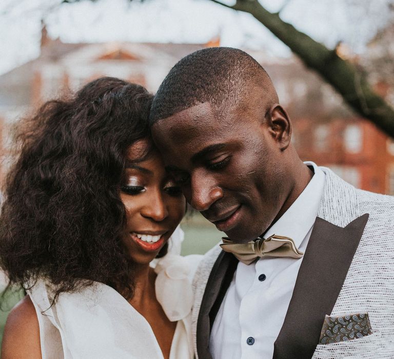 Smiling bride with short curled hair in white satin Bec + Bridge wedding dress with shoulder bows stands holding arm of groom in grey suit jacket with black lapels outside after Bridge Community Church wedding