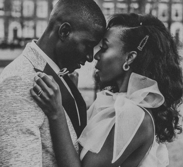 Groom in grey suit jacket with black lapels and bow tie stands with his head touching bride with curled hair and grey checked jacket