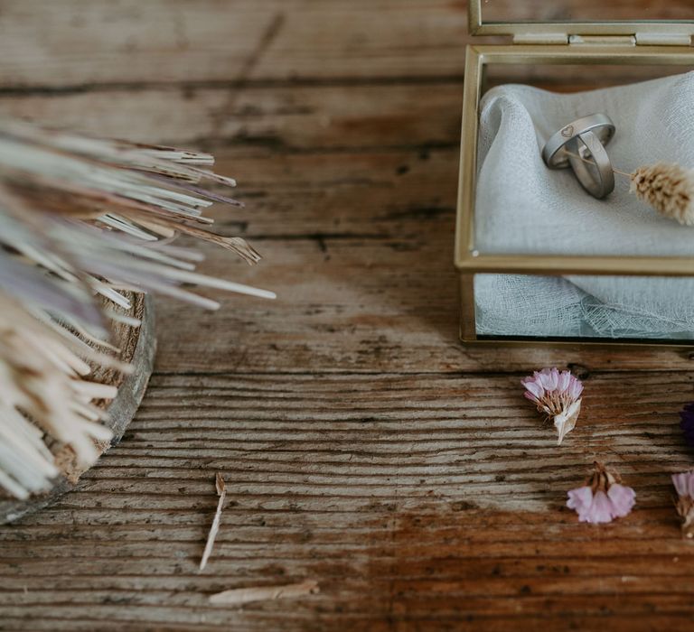 Wedding ring in small gold rimmed glass box on wooden table decorated with dried flowers and ring with dark orange gemstone