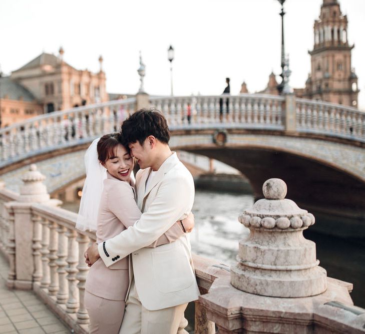 Asian woman wearing veil and pink suit and partner wearing cream suit embracing on Seville riverside