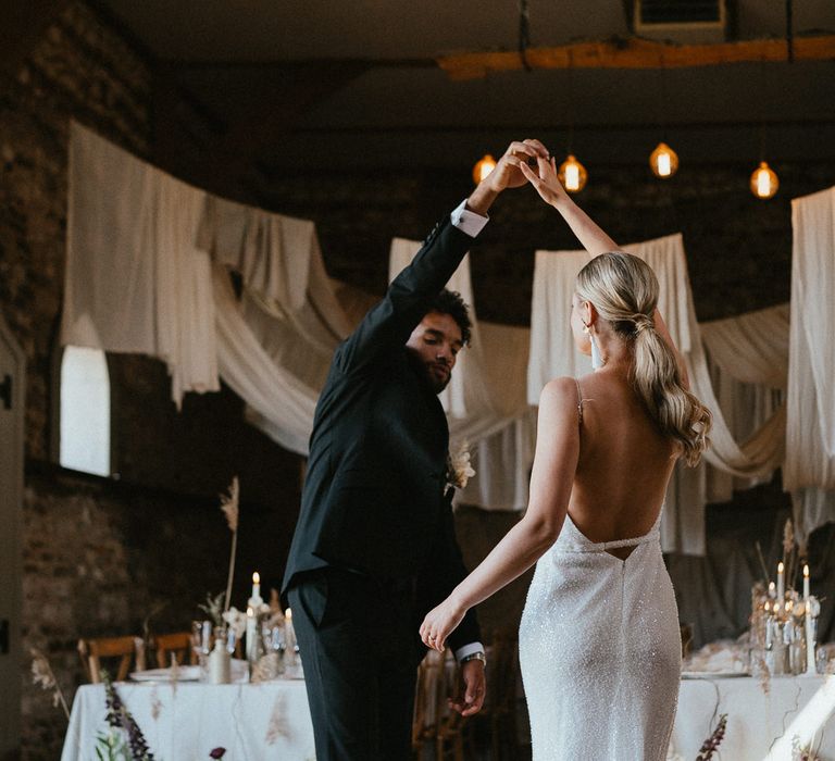 Stylish bride in a low back sparkly wedding dress with sleek ponytail being twirled by her husband in their reception room 