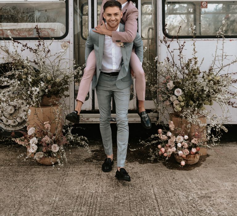 Groom carries groom on his back whilst both wearing pastel suits