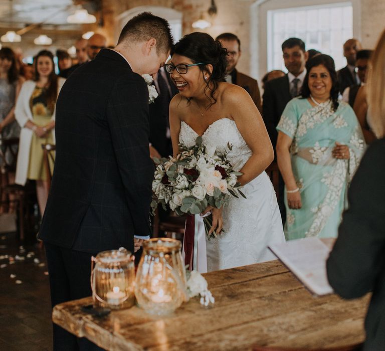 A bride and groom laugh during their wedding ceremony The bride wears glasses and holds a large bouquet of roses, berries and greenery.