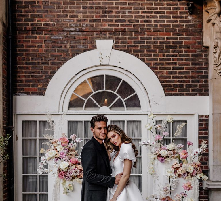 Stylish bride and groom in an all black suit and tie standing at the altar with pink floral arrangements 