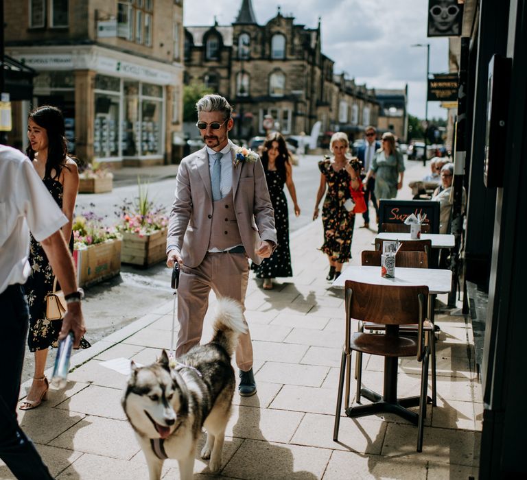 Groom in brown Moss Bros suit, blue tie and sunglasses walks through Harrogate with husky dog before Harrogate wedding