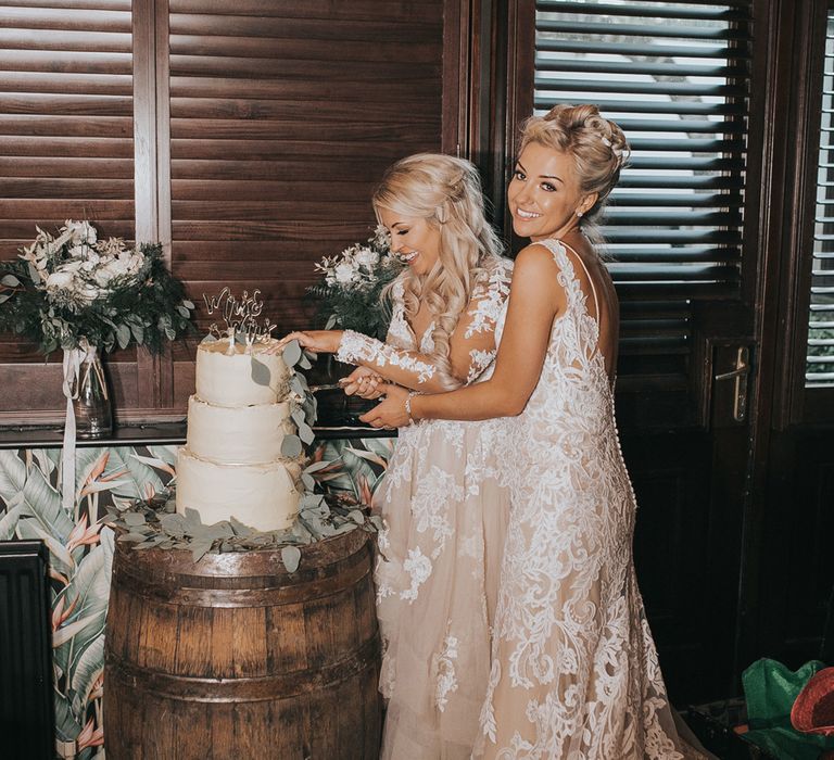 Two brides in white dresses cut a three tiered white wedding cake at Crab & Lobster Fairytale Wedding.