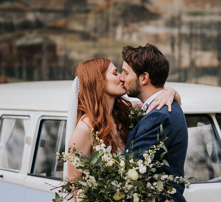 Groom in a navy wool suit kissing his red headed bride in a satin slip dress in front of their VW camper van wedding car 