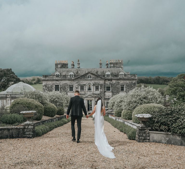 Bride in white Pronovias wedding dress with train and veil walks hand in hand with groom unblock tuxedo towards Came House Dorset